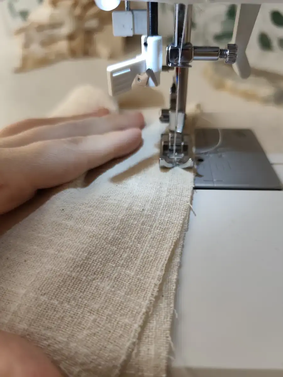 Manos guiando un tejido de algodón crudo bajo el prensatelas de una máquina de coser