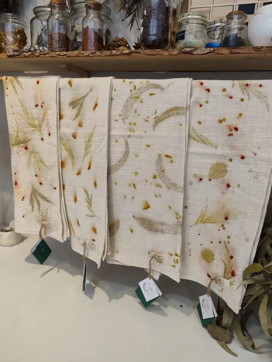 Paños de algodón con estampación botánica de hojas y flores, colgados bajo una estantería con frascos de tintes naturales